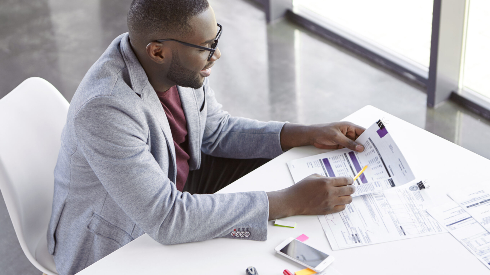 Top view of dark skinned African male copywriter or enterpreneur looks attentively at documents, studies net profit of his company, sits at cozy cabinet, in private office, does paper work alone