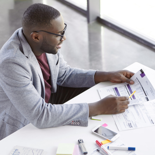 Top view of dark skinned African male copywriter or enterpreneur looks attentively at documents, studies net profit of his company, sits at cozy cabinet, in private office, does paper work alone